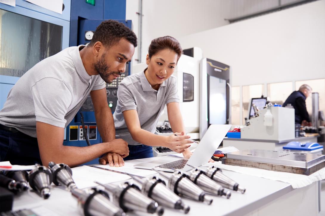 Two people in a manufacturing workshop reviewing information on a laptop, with precision metal machine parts arranged on a table and industrial equipment in the background.